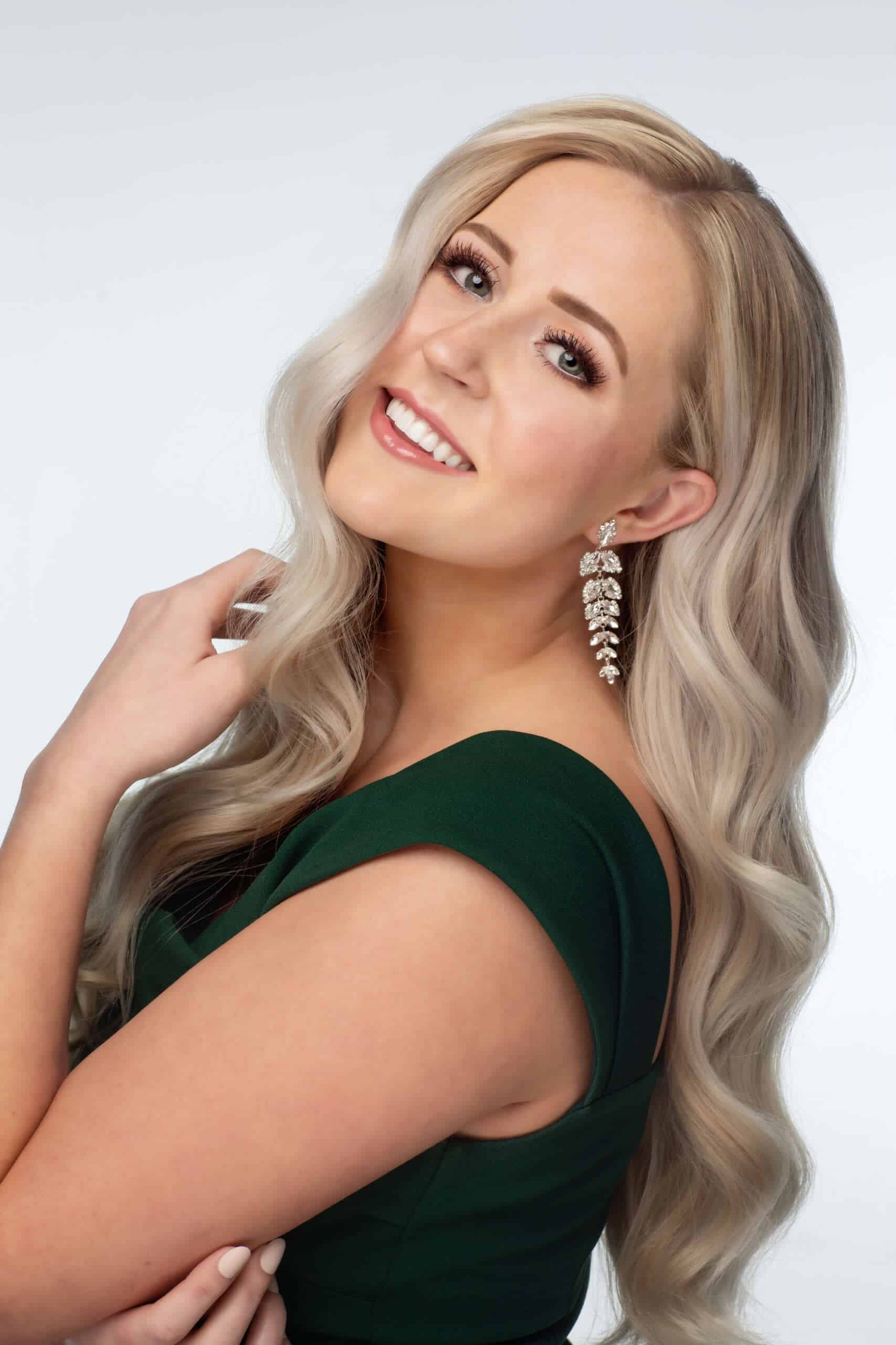 Elegant young woman in formal dress with blonde wavy hair and diamond earrings, smiling for a professional photo on UVU REVIEW website.
