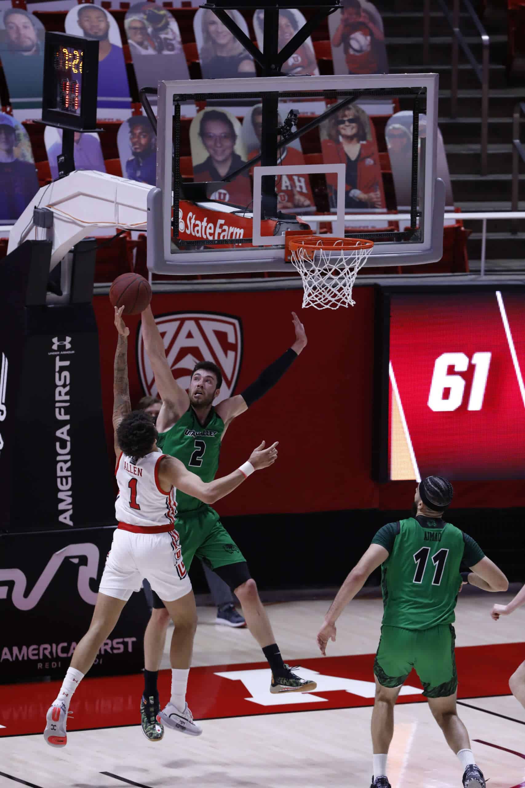 UVU men's basketball game featuring players in action near the hoop, showcasing dynamic gameplay, team uniforms, and arena setting with digital scoreboards and crowd photography in the background.