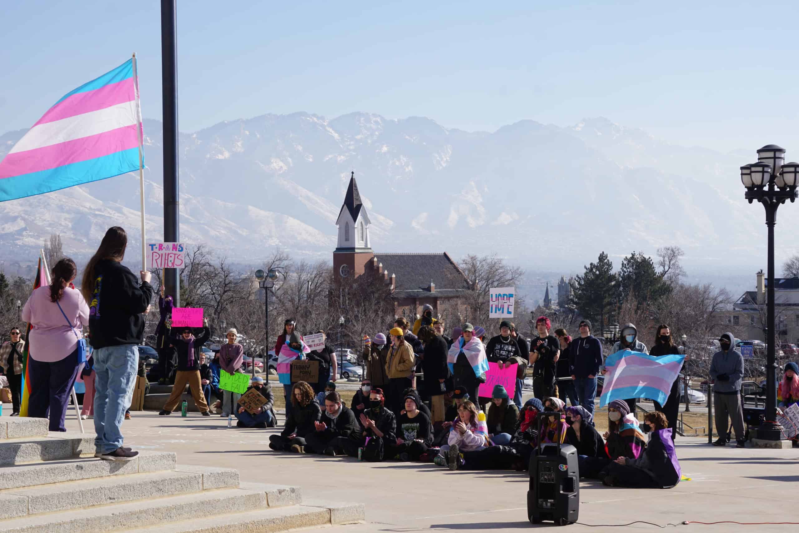 Transgender pride flags and students gathered for a rally at UVU campus with mountains in the background.